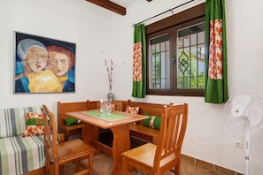 A dining area with a wooden table, chairs, and a bench. There is a painting on the wall, a window with curtains, and a standing fan. - Holiday Home 'Casa Rural La Higuera' with Shared Pool, Wi-Fi and Air Conditioning (Barbate)
