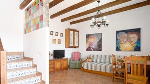 A living room with a staircase, wooden beams, a chandelier, a television, a sofa, a dining table, and a painting.