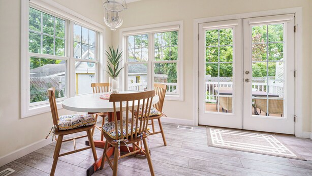 A dining area with a round table, wooden chairs, and large windows.