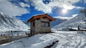 Uma cabana de pedra com telhado de madeira, rodeada por montanhas cobertas de neve e um céu claro.