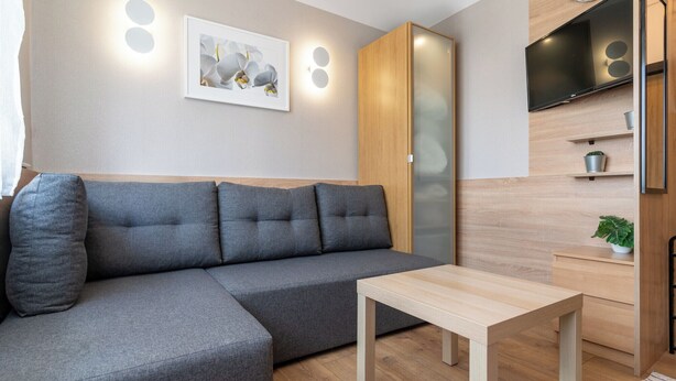 Un soggiorno moderno con un divano angolare grigio, un tavolino in legno, una TV montata a parete e una piccola pianta in vaso su uno scaffale.