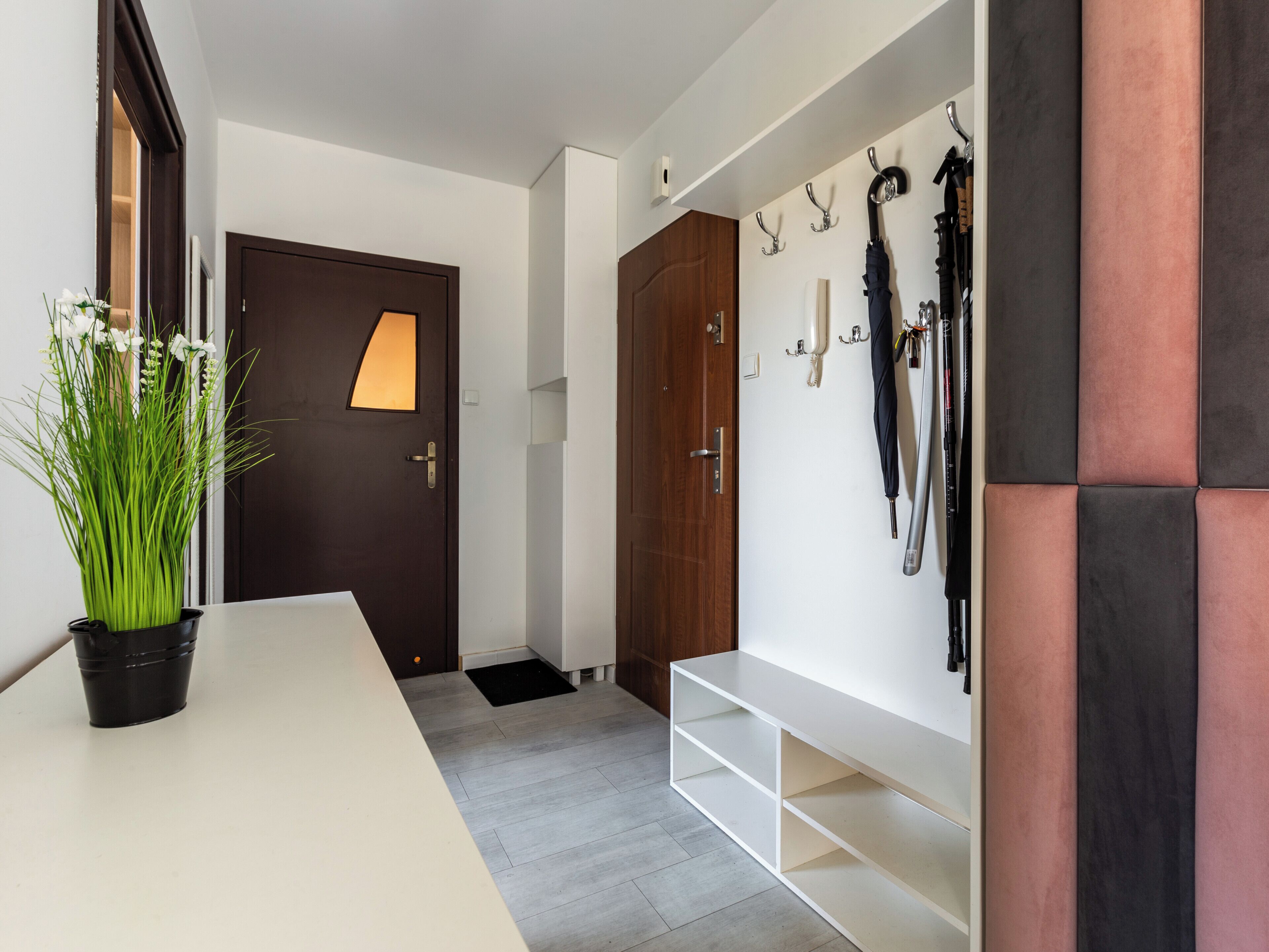 Um corredor com uma receção de cor branca, uma porta de madeira escura, um suporte de guarda-chuvas e um banco branco.
