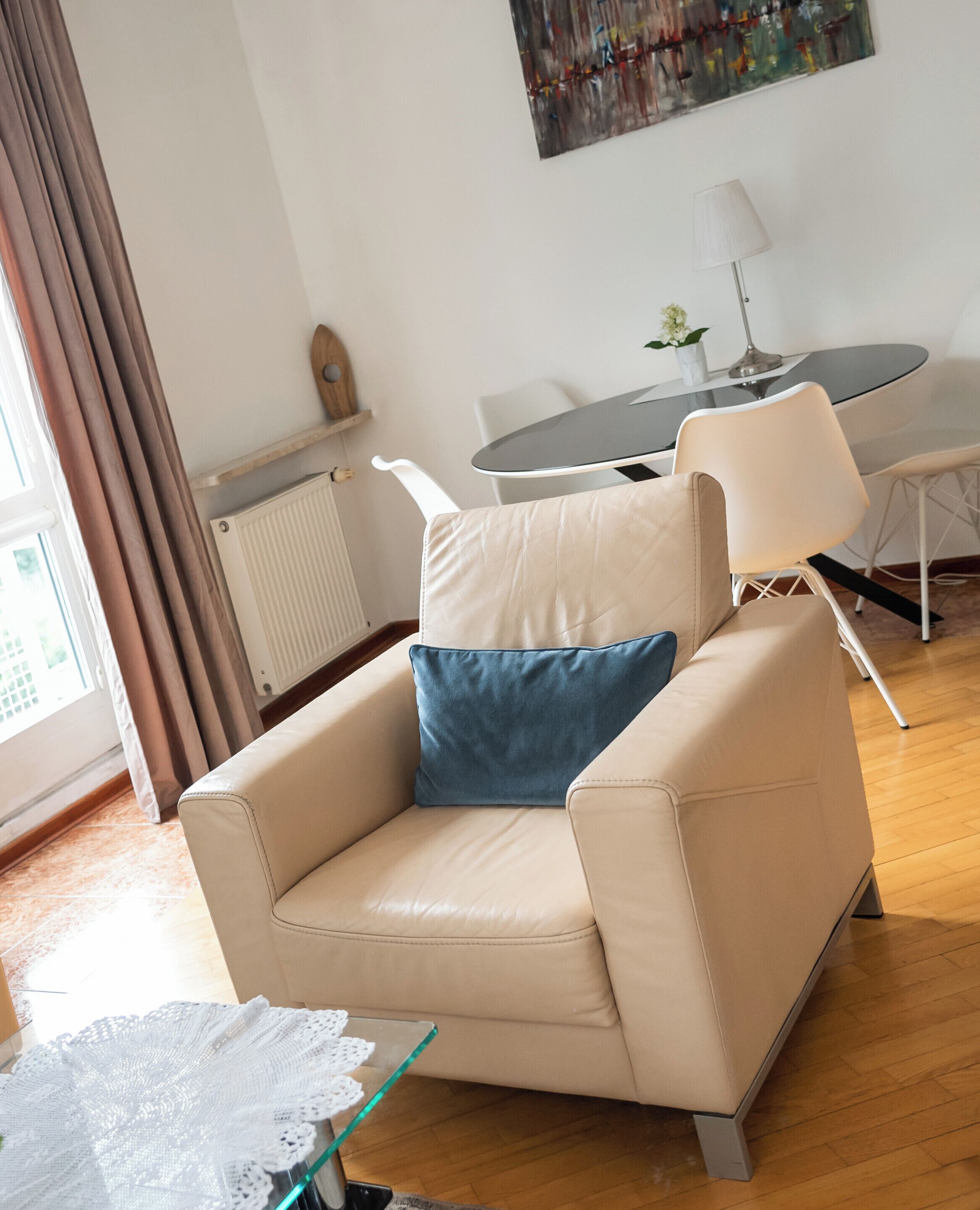 Een beige fauteuil met een blauw kussen, een glazen salontafel met een witte kantplooi, een eettafel met stoelen, een radiator en een raam met gordijnen.