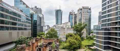 Un paysage urbain avec des immeubles modernes de grande hauteur, un mélange d’architecture en verre et en béton, et une zone centrale où la végétation a envahi les lieux, avec une structure en briques délabrée.