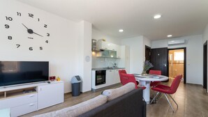 Una sala moderna con un televisor de pantalla plana grande, un sofá, una mesa de comedor con sillas rojas y una cocina con armarios blancos y una estufa.
