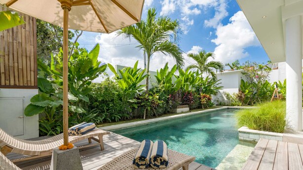 Un espace aménagé au bord de la piscine, avec des chaises longues, un pilier en béton et une piscine entourée d’une végétation luxuriante.