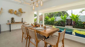 A dining area with a wooden table and woven chairs, a pool view, and decorative wall art. - Villa Palikua (Tabanan)