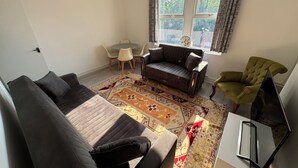En stue med sofa, lænestol, et lille bord med stole og et vindue med gardiner.