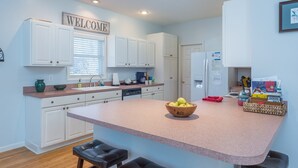 Una cocina moderna con armarios blancos, un letrero de bienvenida, una isla grande con un bol de fruta y un refrigerador.