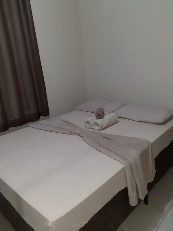 Una cama individual con sábana blanca, manta color beige y una toalla enrollada.