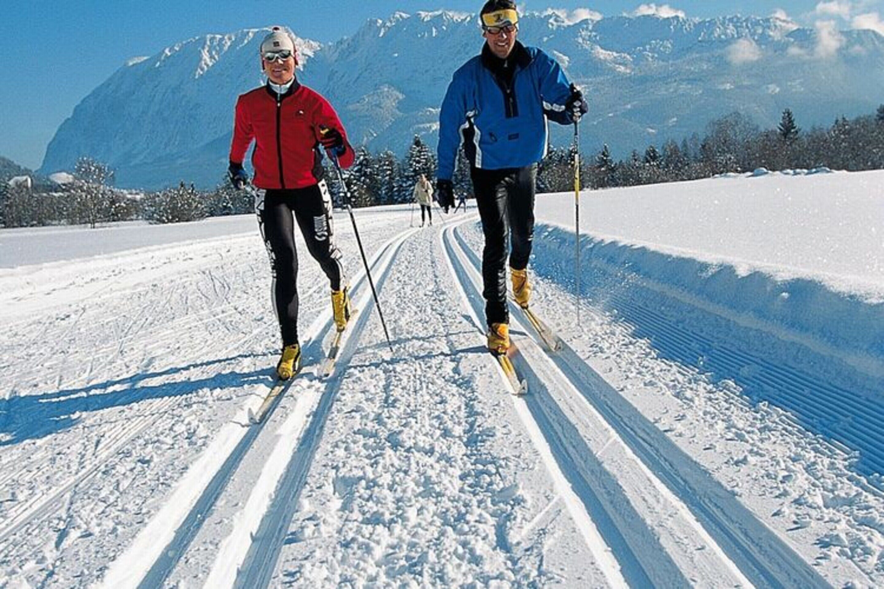 Deux personnes pratiquent le ski de fond sur une piste enneigée, avec une montagne en arrière-plan.