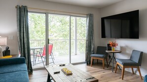 Un salon moderne doté d’une grande fenêtre, d’un téléviseur à écran plat et d’un coin repas comprenant deux chaises et une table.