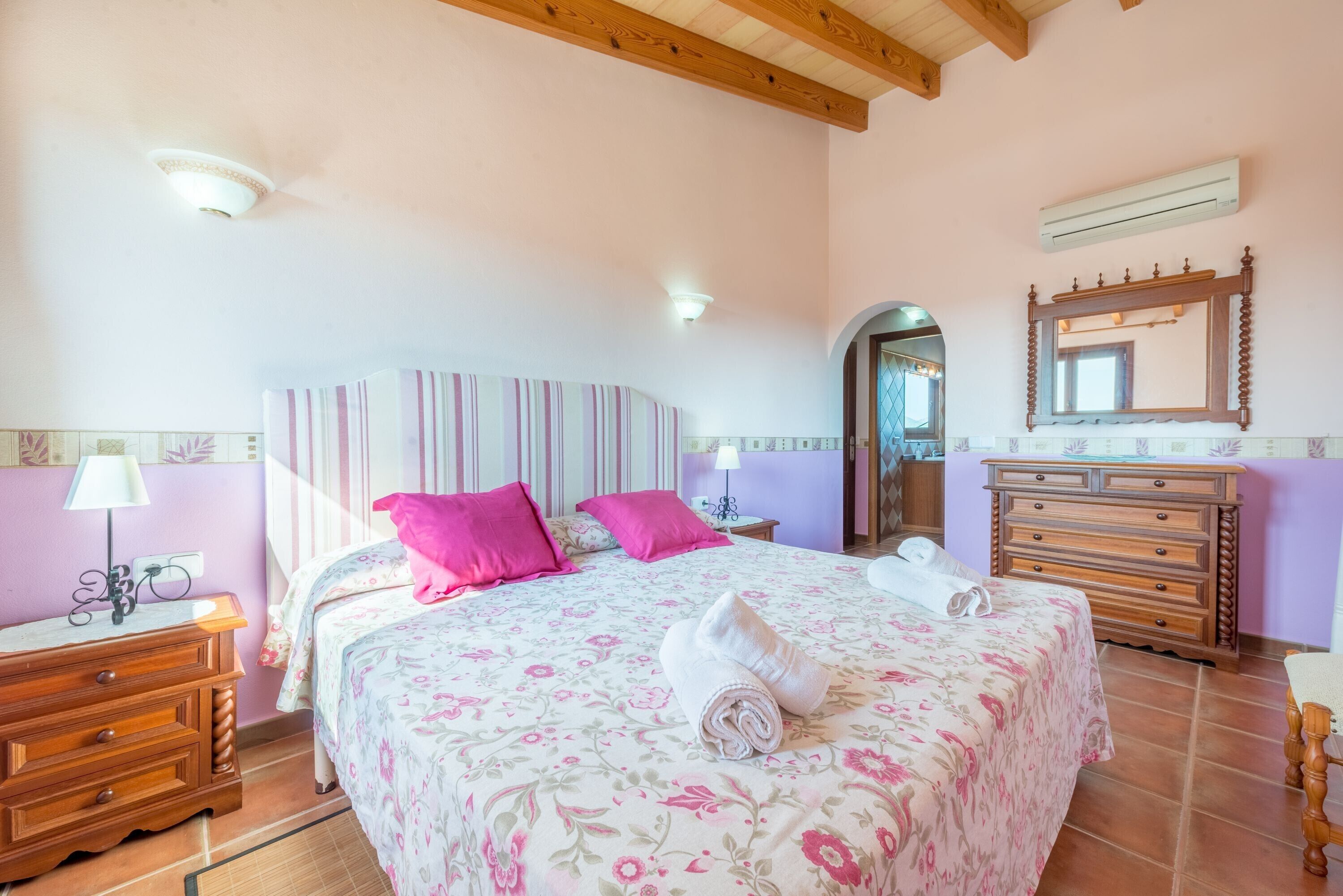 A bedroom with a bed, bedside table, dresser, mirror, and a door leading to another room.