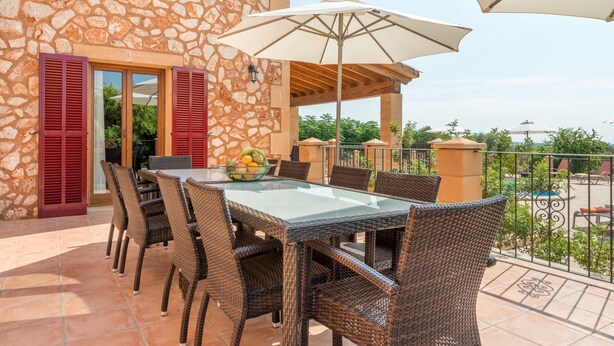 A stone building with a glass table and wicker chairs, a balcony with a railing, and a view of greenery.