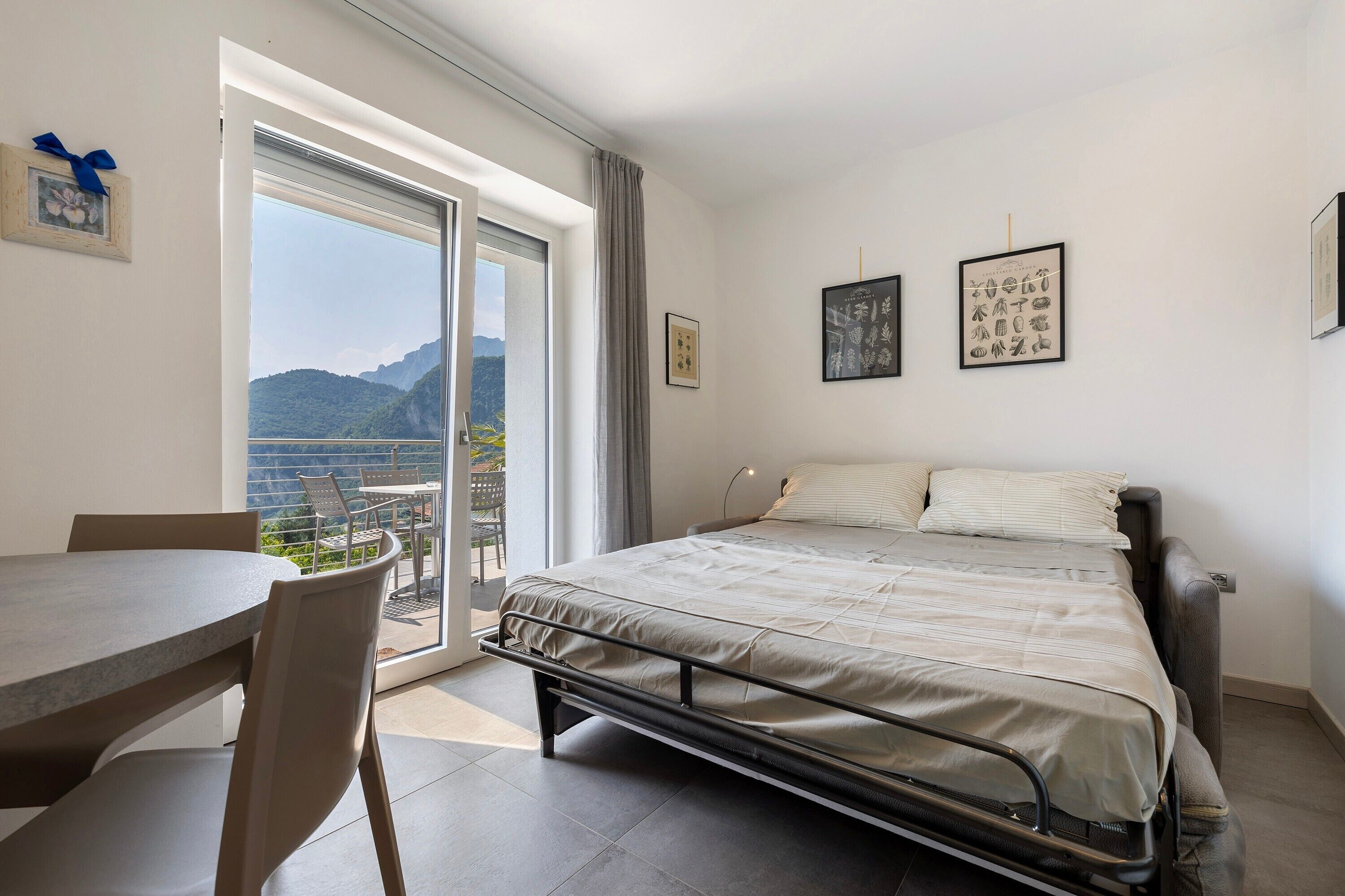 A bedroom with a bed, a table, and chairs, a balcony with a view of mountains, and framed pictures on the wall.