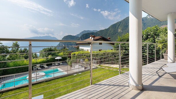 A balcony with a pool, a view of mountains, and a house in the background. - Eco del Garda - Deluxe Studio 101 with Covered Terrace and Lake View (Tenno)