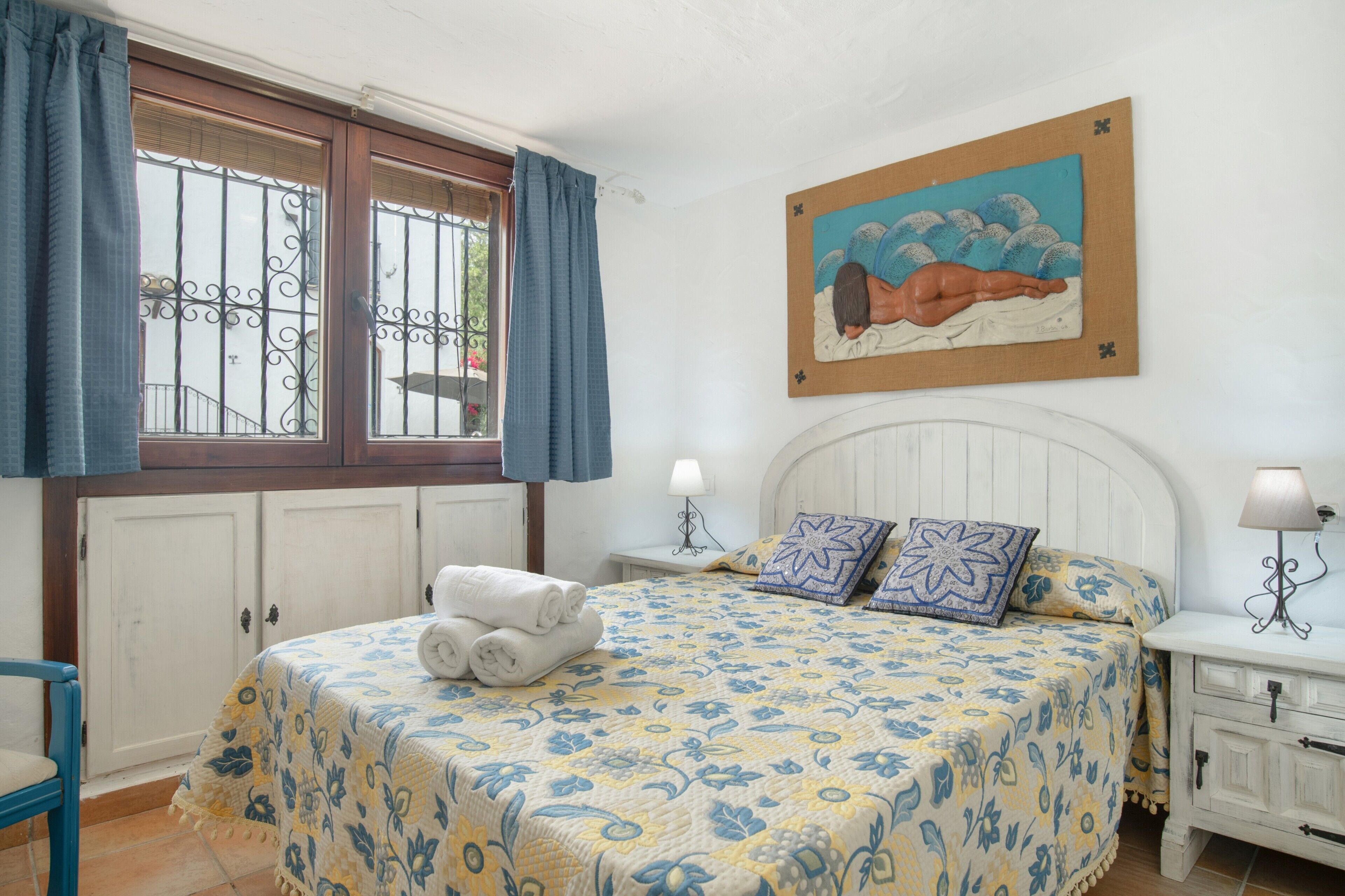 Een slaapkamer met een bed, nachtkastje, lamp, raam met gordijnen en een schilderij aan de muur.