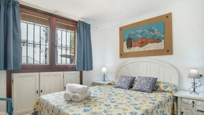 Un dormitorio con cama, mesita de noche, lámpara, ventana con cortinas y un cuadro en la pared.