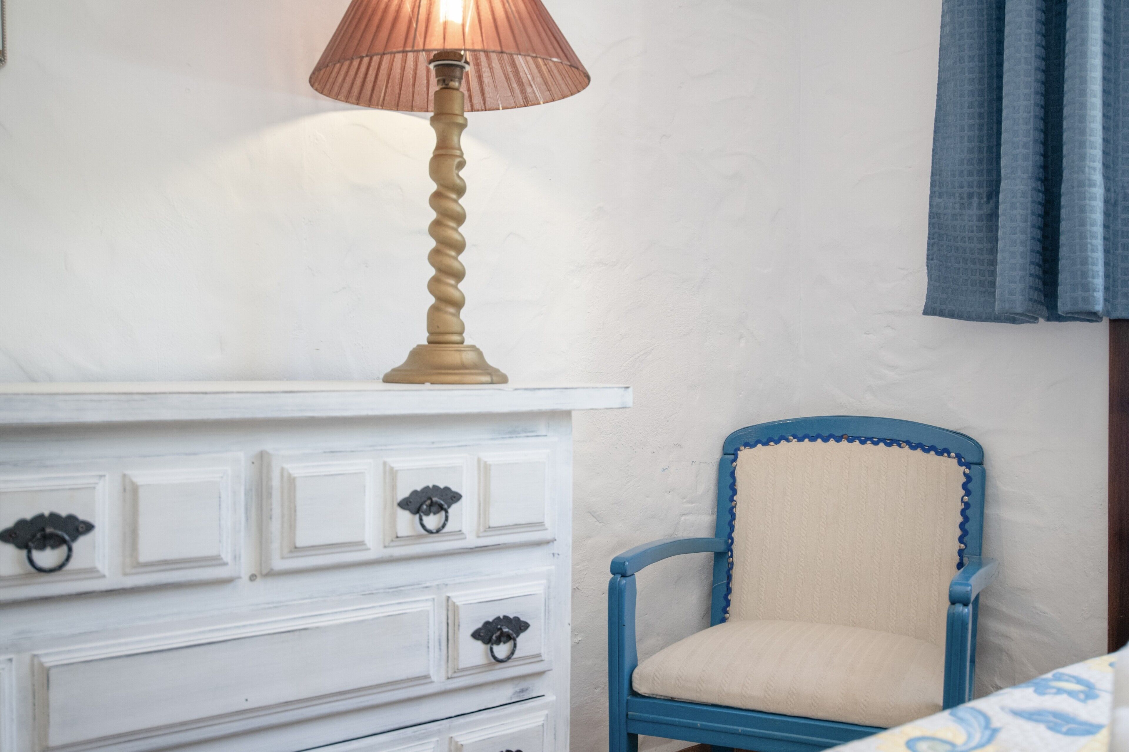 Une commode blanche avec des tiroirs et une lampe, une chaise bleue avec un siège rembourré, et un mur blanc avec une fenêtre.