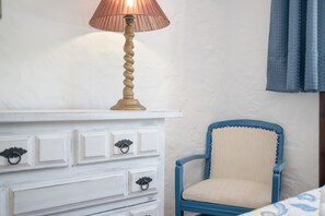 Une commode blanche avec des tiroirs et une lampe, une chaise bleue avec un siège rembourré, et un mur blanc avec une fenêtre.