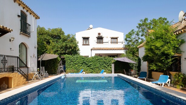 Una piscina con sillas de descanso, un edificio blanco con techo de tejas y un cielo azul despejado.