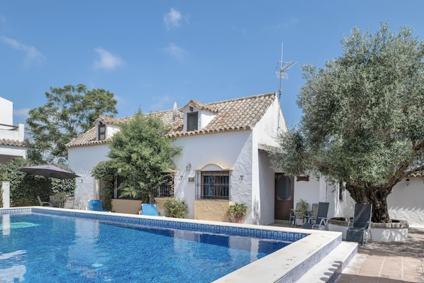Une piscine, un bâtiment blanc avec un toit en tuiles et des oliviers.