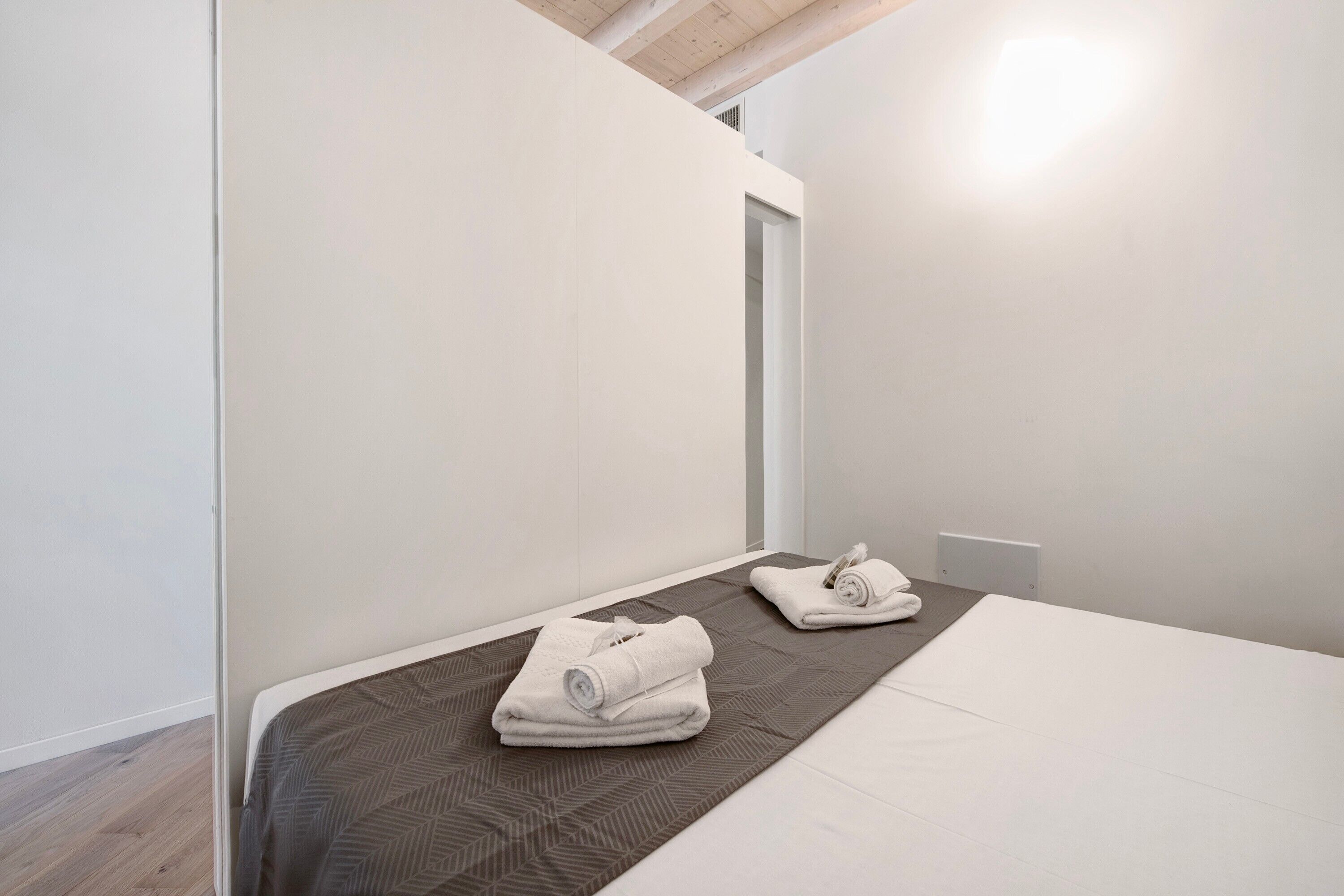 Ein Einzelbett mit brauner, gemusterter Bettdecke, zwei ordentlich gefaltete weiße Handtücher und eine weiße Wand mit einer Tür.