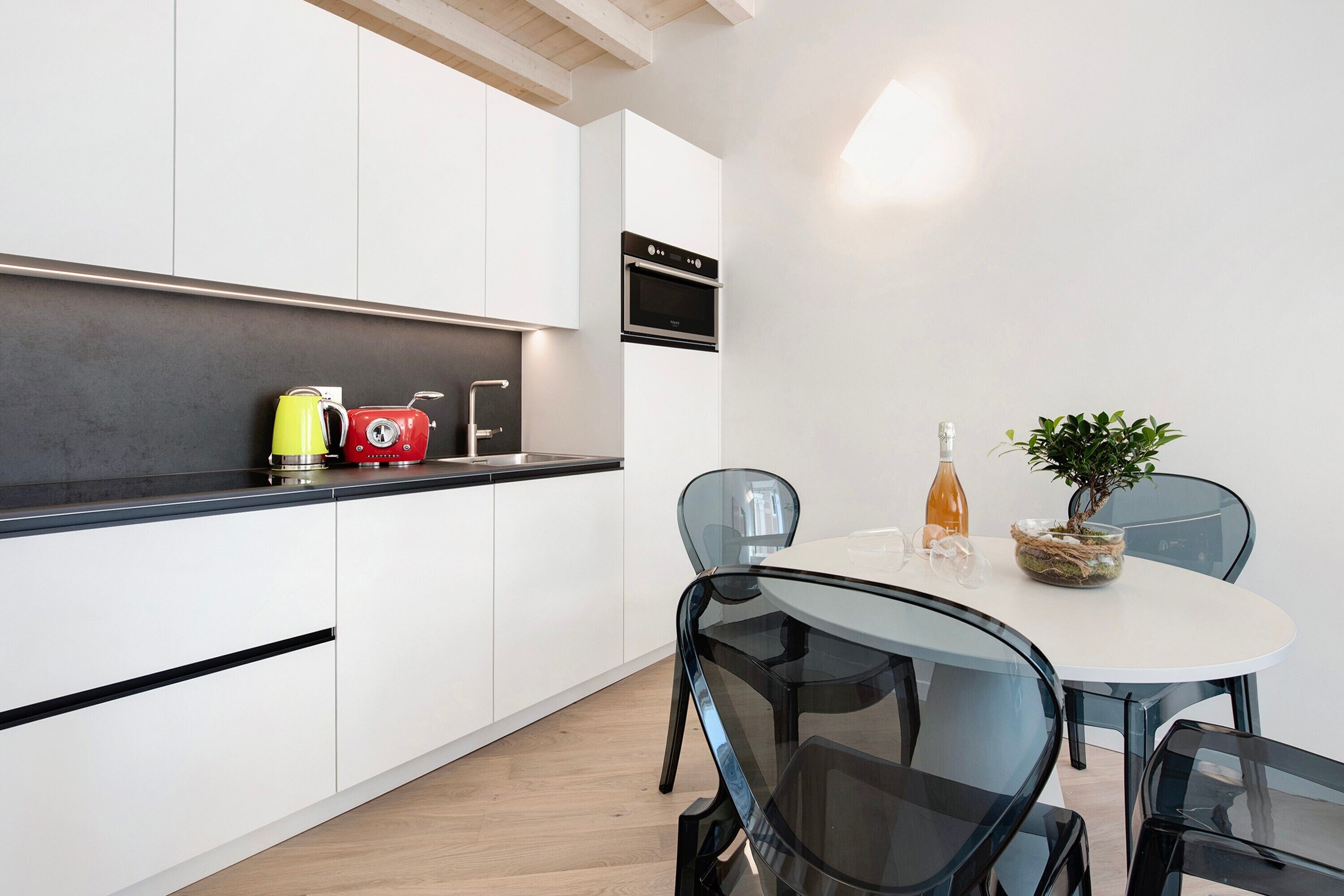 Een moderne keuken met witte kasten, een zwart aanrecht en een inbouwoven. Er is een eettafel met stoelen en een kleine potplant.
