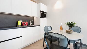 Een moderne keuken met witte kasten, een zwart aanrecht en een inbouwoven. Er is een eettafel met stoelen en een kleine potplant.