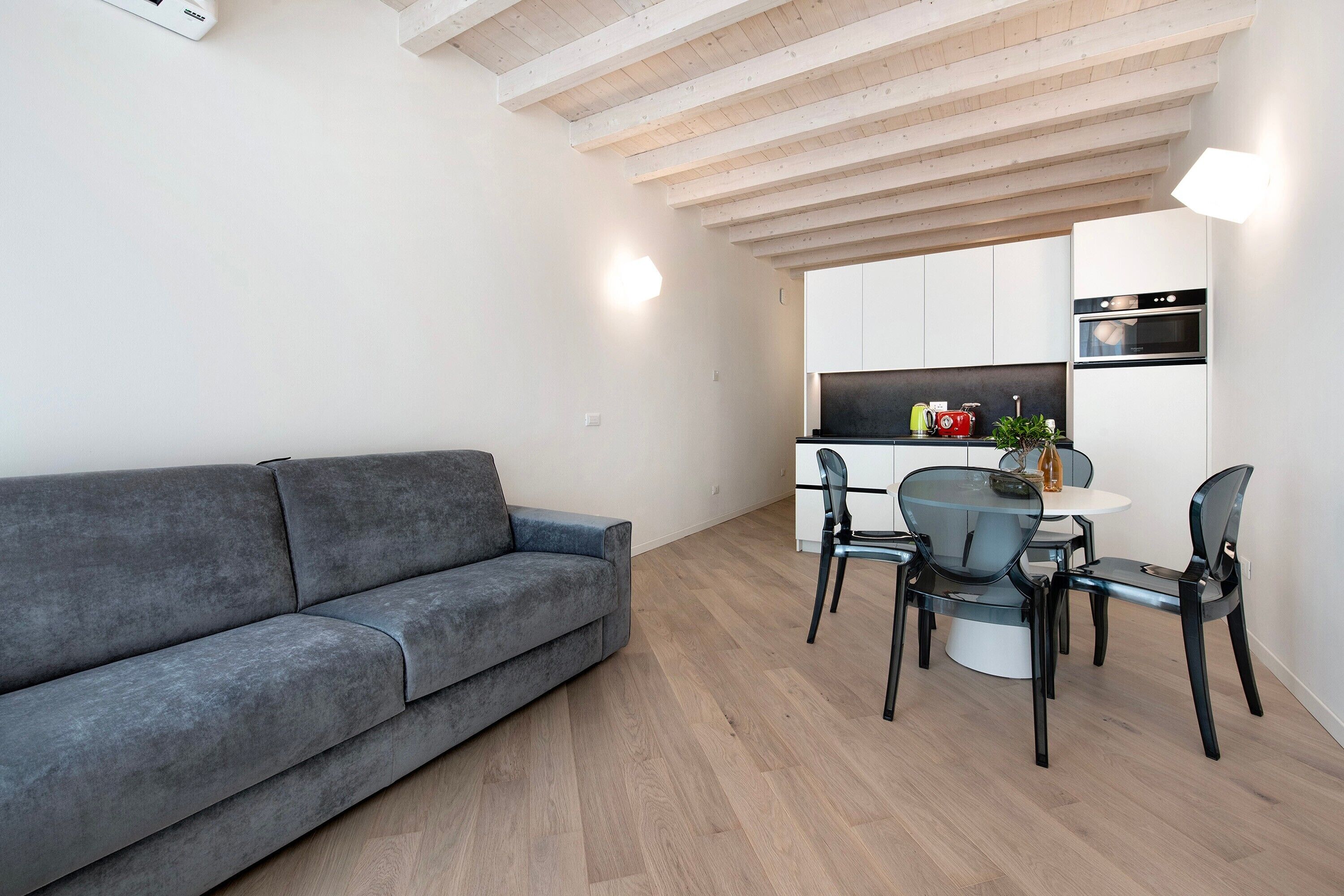 Een moderne woonruimte met een grijze bank, een eettafel met stoelen, een keuken met kasten en apparatuur, en een houten plafond.