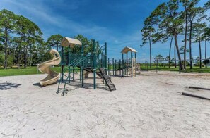 Ein Spielplatz mit Rutsche und Klettergerüsten, umgeben von Sand und Bäumen.