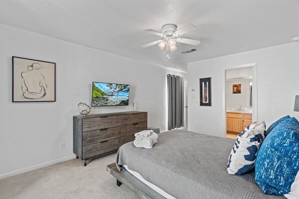 A bedroom with a bed, a dresser, a ceiling fan, a TV, and a bathroom visible through an open door.