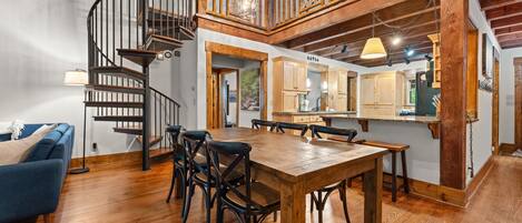 A spacious dining area with a wooden table and chairs, a spiral staircase, and a kitchen area with wooden cabinets and a hanging light fixture.