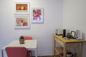 Ein Zimmer mit einem Esstisch, Stühlen, einer kleinen Topfpflanze, einem Holznachttisch mit Toaster, Wasserkocher und einer Weinflasche.
