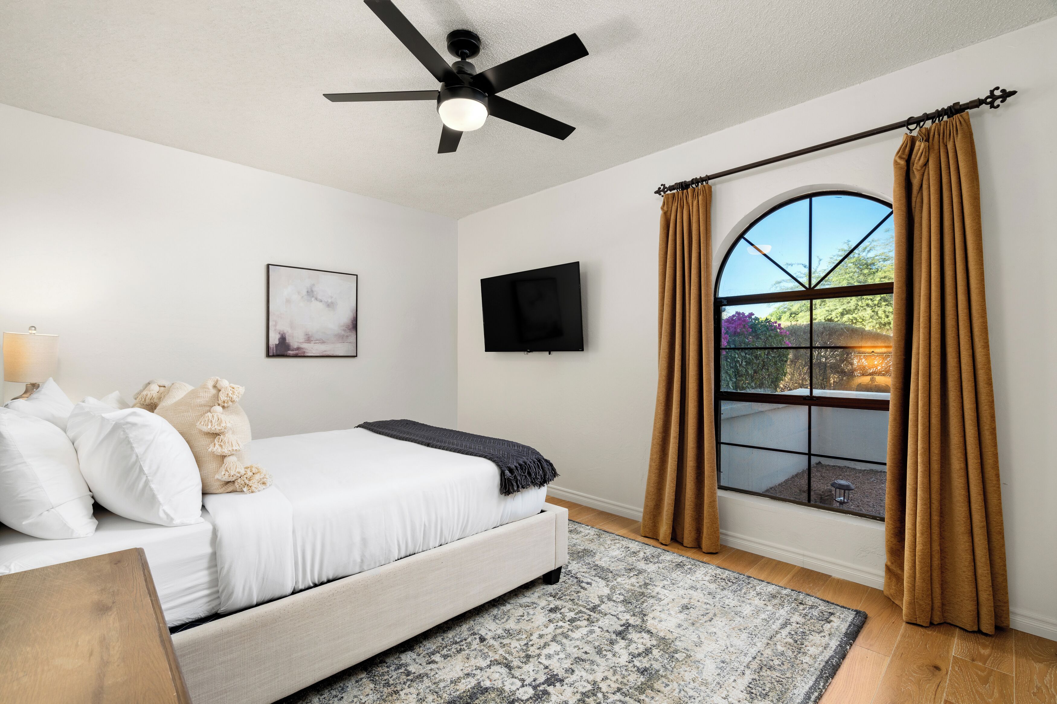 Une chambre avec un lit, un ventilateur de plafond, une télévision, une fenêtre avec des rideaux et une vue sur la verdure à l’extérieur.