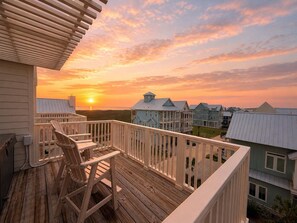 Una terraza de madera con sillas, vistas a casas y un atardecer.