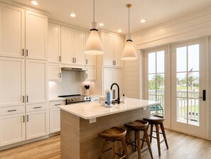Una cocina con armarios blancos, una isla central con taburetes, iluminación colgante y vistas al exterior a través de grandes ventanales.