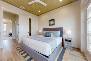 Un dormitorio con cama, mesitas de noche, una mesita auxiliar, ventilador de techo, ventana y puerta que da a otra habitación.