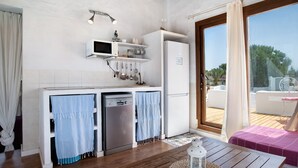 A compact kitchen with a microwave, oven, and dishwasher. There is a wooden table with a lantern and a pink chair. A sliding glass door leads to an outdoor area with a wooden deck and a white chair.