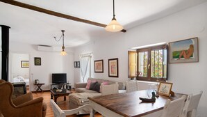 A living room with a wooden dining table, white chairs, a brown armchair, a sofa with cushions, a television, and framed artwork on the walls.