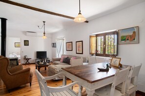 Un salon comprenant une table à manger en bois, des chaises blanches, un fauteuil marron, un canapé avec des coussins, un téléviseur et des œuvres d’art encadrées accrochées aux murs.