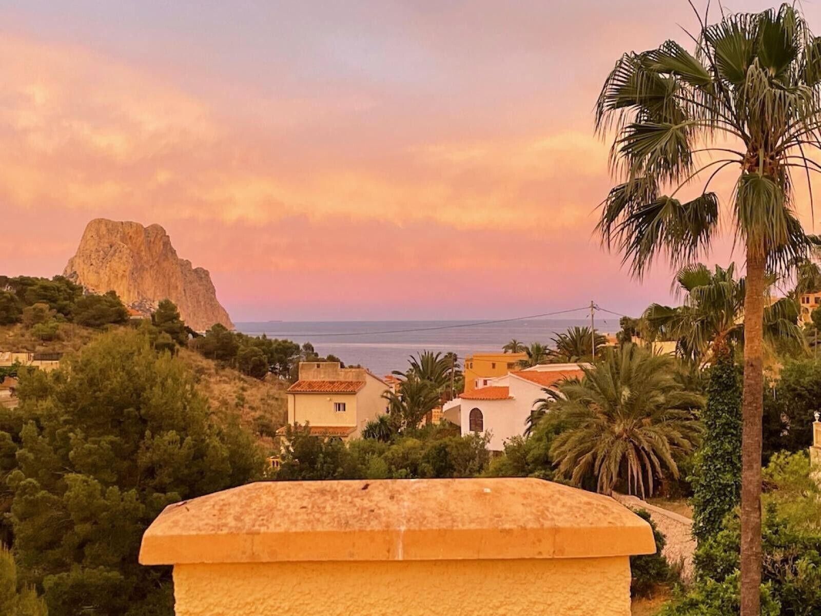 A coastal view with a prominent rock formation, houses with red-tiled roofs, and palm trees.