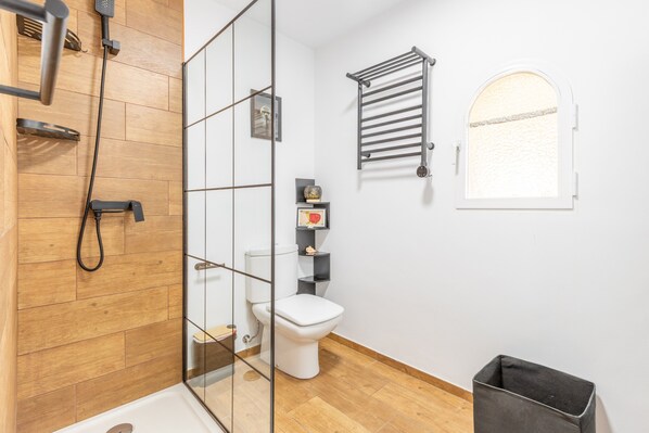 A bathroom with a glass shower enclosure, a white toilet, a towel rack, and a small window.