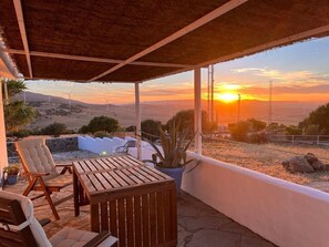 Un patio al aire libre con una mesa, sillas, una planta en maceta y vistas a aerogeneradores y al atardecer.