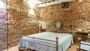 Quarto com parede de tijolos, estrutura de cama de metal, mesa de cabeceira com abajur e uma pequena prateleira.