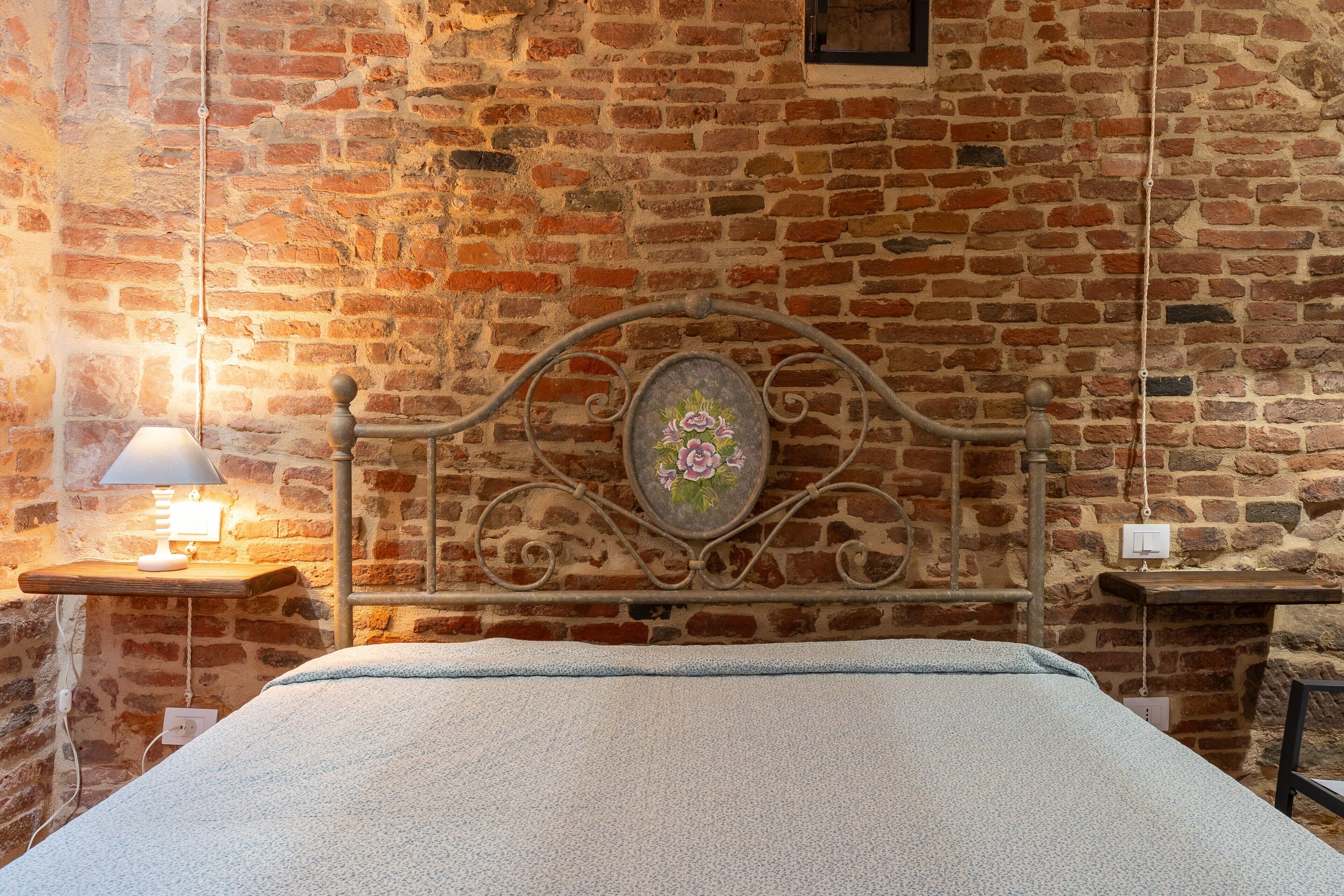 Une pièce avec un mur en briques, un cadre de lit en métal, une table de chevet avec une lampe et une petite étagère.