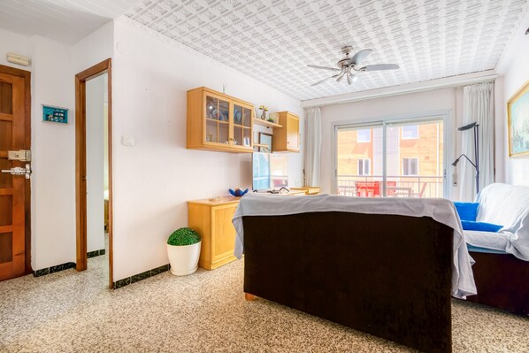Une pièce comprenant un lit, un bureau, une armoire, une plante en pot et une fenêtre donnant sur des immeubles.