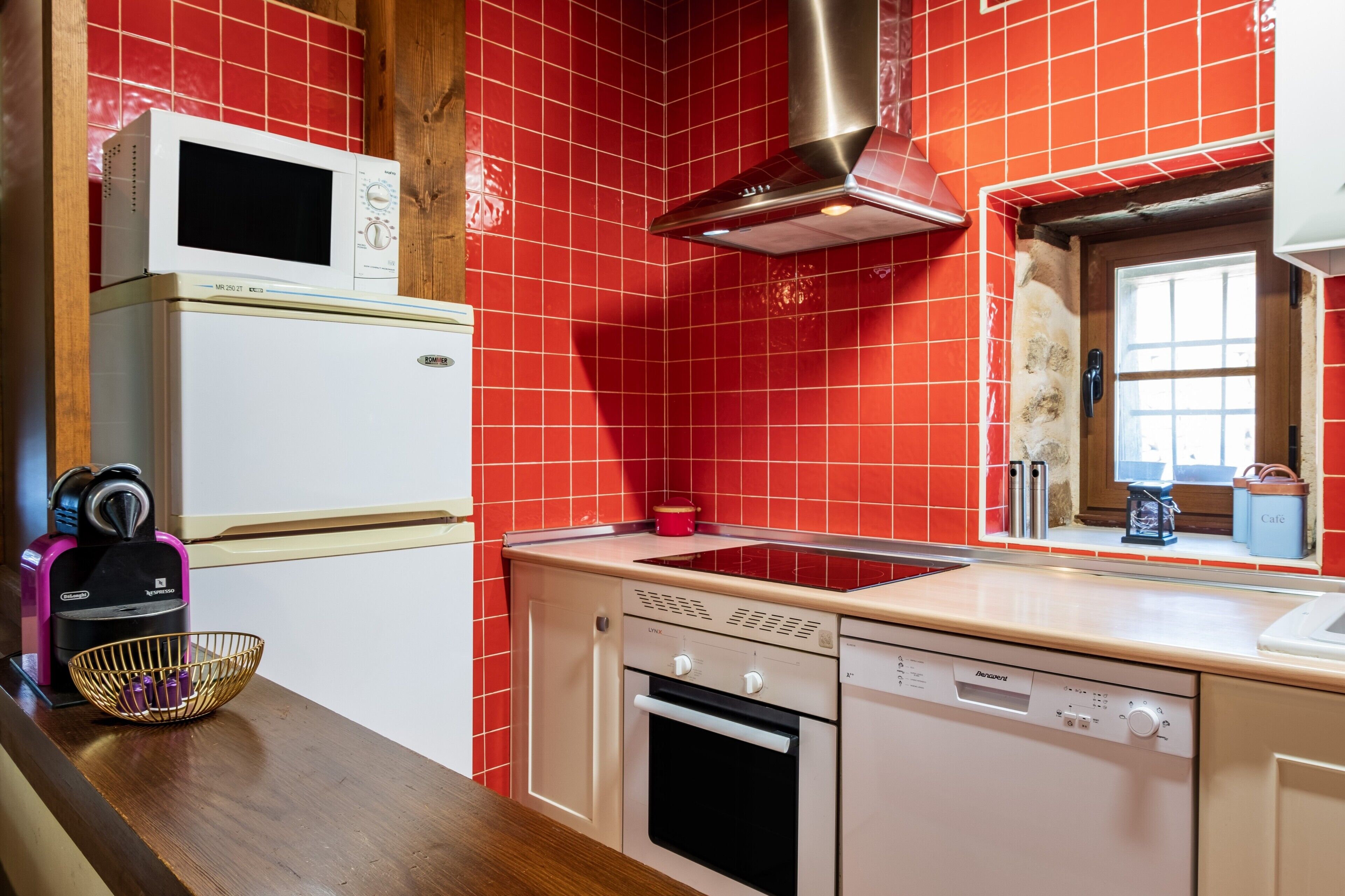 Una cocina compacta equipada con microondas, horno y lavavajillas, una encimera de madera y paredes con azulejos rojos.