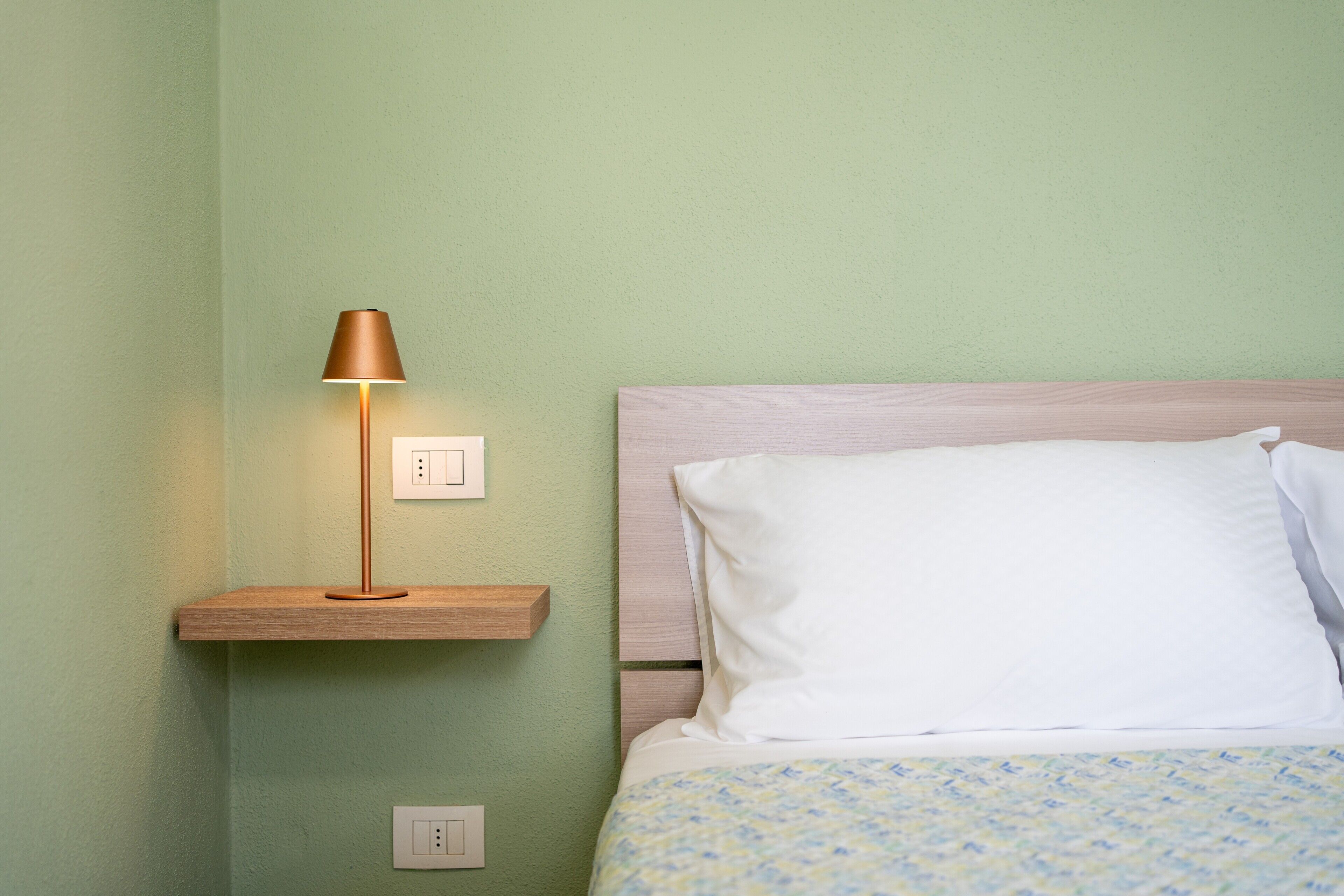 Un lit avec un oreiller blanc et une couverture à motifs, une lampe de chevet posée sur une étagère en bois, et un interrupteur sur le mur.