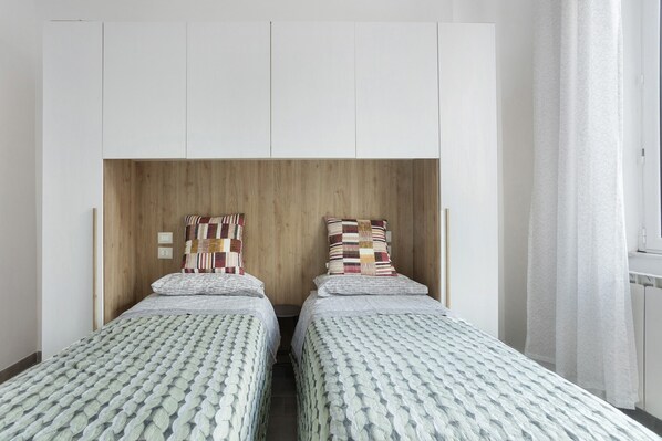 A room with two beds, a wooden headboard, white wardrobe, and a window with curtains. - Apartment 'Georgehouse Golfo Dei Poeti 5 Terre' with Wi-Fi (La Spezia)